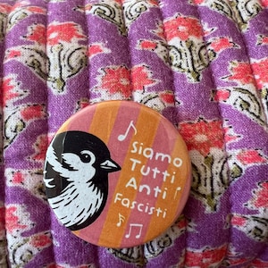 May include: A round button with a black and white bird illustration on a striped orange and pink background. The button has the text "Siamo Tutti Anti Fascisti" and musical notes. The button is on a purple floral patterned fabric.