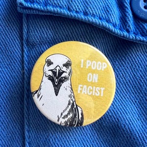 Peut inclure: Un bouton rond et jaune avec une illustration en noir et blanc d'une tête d'oiseau. Le bouton porte le texte "I POOP ON FACIST" en blanc. Le bouton est épinglé sur une veste en jean bleue.