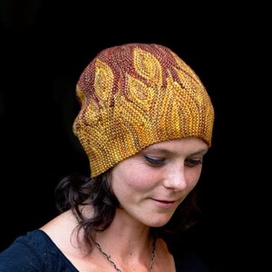 May include: A knitted beanie in shades of yellow and brown, featuring a leaf-like pattern. The hat is worn by a person wearing a dark blue top and a silver necklace. The background is a solid black.