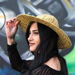 Peut inclure: Un chapeau de soleil de couleur paille avec un large bord. Le chapeau est maintenu. La personne porte un chemisier noir à manches longues. Le fond est un mur de graffitis colorés.