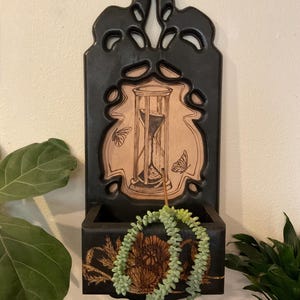 May include: Black wooden wall planter with an hourglass and butterfly design. The planter has a small box for plants and a decorative top. A green succulent plant is in the planter.