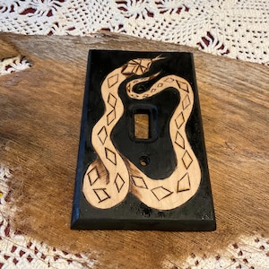 May include: A black light switch cover with a carved snake design. The snake is light brown with diamond-shaped markings. The switch plate has a rectangular shape with a single switch opening and two screw holes.