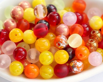 May include: A collection of colorful, round beads in various shades of yellow, orange, red, and pink. Some beads have a translucent or frosted appearance, while others are opaque. The beads are displayed in a white, shallow dish.