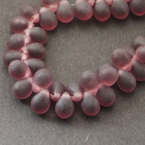 Op de afbeelding: Een streng van matte, druppelvormige kralen in mauve en roze tinten. De kralen zijn aan elkaar geregen, wat een delicate uitstraling creëert. De kralen zijn waarschijnlijk gemaakt van glas of een vergelijkbaar materiaal.