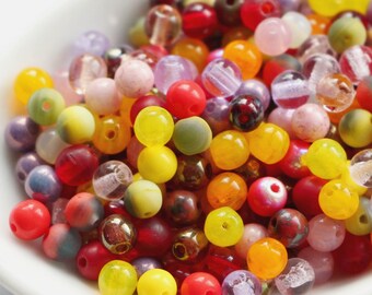 May include: A collection of colorful beads in various sizes and textures, including translucent, matte, and glossy finishes. The beads are in shades of red, yellow, pink, purple, and clear, displayed in a white, shallow dish. Ideal for jewelry making.
