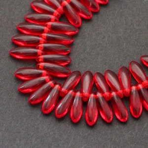Könnte beinhalten: Eine Kette aus durchscheinenden, roten, tropfenförmigen Perlen. Die Perlen sind mit einem roten Faden aufgereiht und bilden eine gebogene Form. Die Perlen sind gleichmäßig verteilt und haben eine glänzende Oberfläche. Der Hintergrund ist eine dunkelgraue Oberfläche.