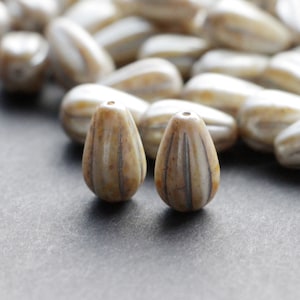 May include: Close-up of a collection of teardrop-shaped beads. The beads are a light beige color with vertical brown stripes. The beads have a glossy finish and are scattered on a dark gray surface. These beads are ideal for jewelry making.