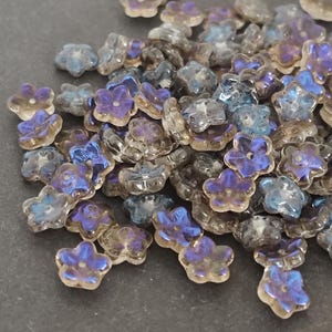 May include: A collection of iridescent flower-shaped beads in shades of blue, purple, and clear. The beads are scattered, showcasing their delicate floral design and shimmering surfaces. These beads are ideal for jewelry making and craft projects.