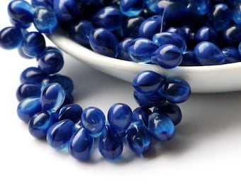 May include: A collection of deep blue, teardrop-shaped glass beads. The beads are arranged in a curved line, with some spilling out of a white bowl. The beads have a glossy finish and are strung together.