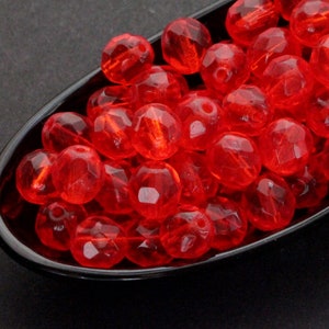 Op de afbeelding: Een verzameling gefacetteerde, transparante rode kralen. De kralen zijn rond met meerdere platte oppervlakken, waardoor een sprankelend effect ontstaat. Ze liggen in een zwarte, ovaalvormige schaal tegen een donkere achtergrond. Ideaal voor het maken van sieraden of knutselprojecten.