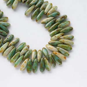 May include: A necklace featuring elongated, teardrop-shaped beads. The beads are predominantly green and white with brown detailing, arranged in a curved design against a white backdrop. The beads have a glossy finish and are strung together.