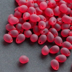 May include: A pile of small, red, teardrop-shaped beads with a frosted finish. Each bead has a small hole at the top for threading. The beads are scattered on a dark grey surface, creating a textured appearance.