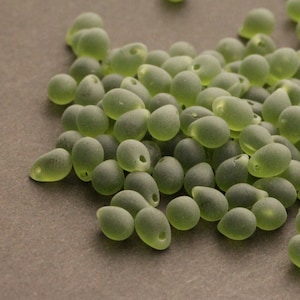 May include: A collection of frosted, olive-green glass beads. Each bead is teardrop-shaped with a small hole at the top for stringing. The beads have a matte finish and are scattered across a neutral surface.