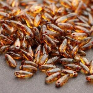 May include: A collection of amber-colored, teardrop-shaped glass beads. Each bead has a small hole at the top and a slightly darker hue towards the bottom. The beads are scattered across a gray surface, creating a textured appearance.