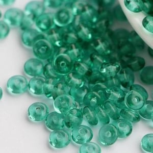 May include: A close-up of teal glass beads, scattered on a white surface and spilling from a white bowl. The beads are small, round, and have a hole through the centre. The beads are a vibrant teal colour, perfect for crafting.