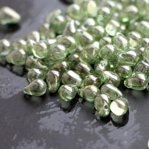 Könnte beinhalten: Eine Ansammlung kleiner, tropfenförmiger Glasperlen in einem blassen Grünton. Die Perlen sind auf einer dunklen, reflektierenden Oberfläche verstreut, wodurch ein Spiel aus Licht und Schatten entsteht. Die durchscheinenden Perlen haben eine glatte, abgerundete Form.