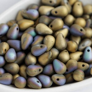 Op de afbeelding: Een verzameling druppelvormige kralen in een witte kom. De kralen hebben een matte afwerking met een kleurengradiënt van goud, blauw en groen. Elke kraal heeft een klein gaatje om te rijgen.