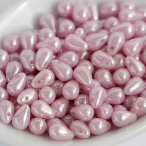 Op de afbeelding: Een verzameling parelmoer roze druppelvormige kralen. De kralen hebben een glanzende afwerking en een klein gaatje om te rijgen. De kralen liggen in een witte kom. Ideaal voor het maken van sieraden.