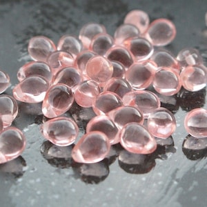 Op de afbeelding: Een verzameling kleine, doorschijnende, druppelvormige kralen in een zachte roze tint. De kralen zijn verspreid over een donker, reflecterend oppervlak, waardoor een spel van licht en schaduw ontstaat. Deze kralen kunnen worden gebruikt voor het maken van sieraden of andere knutselprojecten.