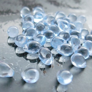 Op de afbeelding: Een verzameling kleine, lichtblauwe, druppelvormige glazen kralen. De kralen zijn verspreid over een reflecterend, donker oppervlak, waardoor een spel van licht en schaduw ontstaat. De doorschijnende kralen hebben een gladde, afgeronde uitstraling.