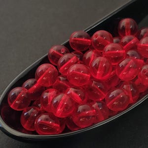 Puede incluir: Un conjunto de cuentas rojas transparentes con un agujero central, presentadas en una cuchara negra brillante. Las cuentas esféricas son probablemente de vidrio o un material similar. La imagen está bien iluminada, destacando la claridad de las cuentas.