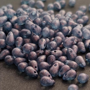 Op de afbeelding: Een stapel druppelvormige kralen in blauwe en paarse tinten. De kralen hebben een gespikkelde, getextureerde uitstraling en een gat om te rijgen. Geschikt voor het maken van sieraden en knutselwerk.