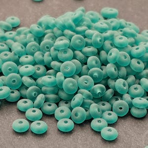 May include: A pile of opaque, teal-colored, donut-shaped beads. The beads are uniform in size and have a matte finish. They are scattered across a neutral-toned surface, suggesting a craft or jewelry-making theme.