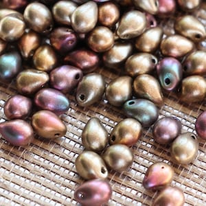 Op de afbeelding: Een verzameling metallic druppelvormige kralen in goud, brons en iriserende paarse tinten. Elke kraal heeft een klein gaatje om te rijgen. De kralen liggen verspreid op een getextureerd, lichtbruin oppervlak.