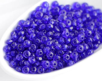 May include: A collection of small, faceted, translucent cobalt blue glass beads. Each bead features a central hole and a multi-faceted surface that reflects light, creating a sparkling appearance across the pile of loose, round-shaped components.
