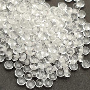 May include: A close-up of a collection of small, clear, round plastic buttons. Each button has a slightly domed shape with a small hole in the centre. The buttons are scattered on a dark surface.