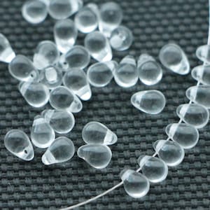 May include: Transparent, teardrop-shaped glass beads, some strung on a white thread, others scattered. The beads have a smooth, rounded form with a small hole at the top for stringing. The background is a dark, textured surface.