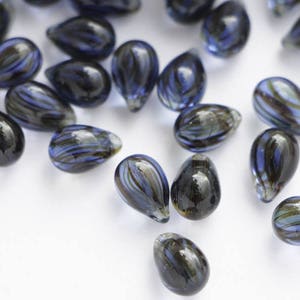 Pu&ograve; includere: Una collezione di perline di vetro a forma di goccia. Le perline sono principalmente blu scuro con striature di blu chiaro, verde e marrone. Sono sparse su una superficie bianca, evidenziando la loro trasparenza.
