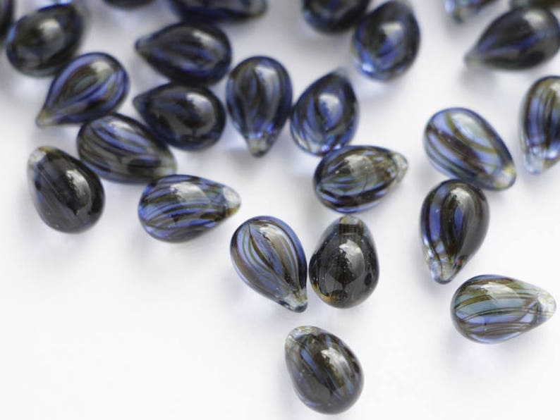 Pu&ograve; includere: Una collezione di perline di vetro a forma di goccia. Le perline sono principalmente blu scuro con striature di blu chiaro, verde e marrone. Sono sparse su una superficie bianca, evidenziando la loro trasparenza.