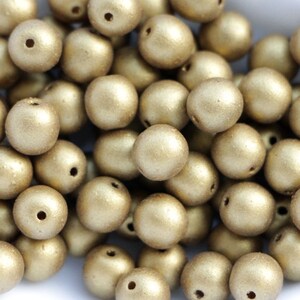 May include: A pile of round, gold-coloured beads with a matte finish. Each bead has a small hole, suggesting they are for stringing. The beads are scattered, creating a textured appearance. Suitable for jewellery making or craft projects.