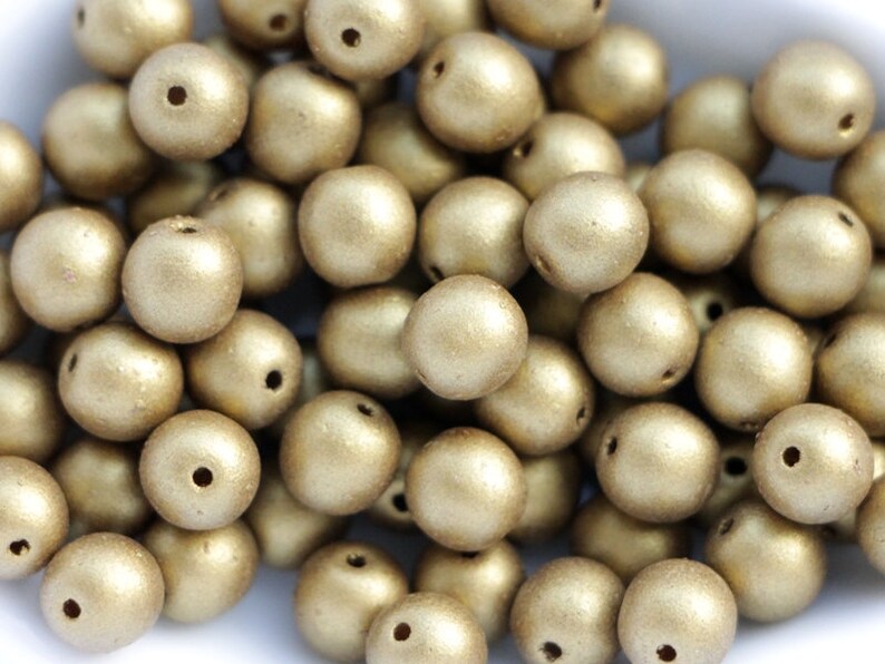 May include: A pile of round, gold-coloured beads with a matte finish. Each bead has a small hole, suggesting they are for stringing. The beads are scattered, creating a textured appearance. Suitable for jewellery making or craft projects.