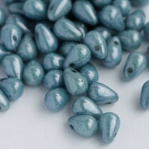 Op de afbeelding: Een close-up van talrijke, traanvormige kralen in teal. Elke kraal heeft een glanzende afwerking en een klein gaatje om te rijgen. De kralen zijn verspreid over een witte ondergrond, wat een textuurrijke weergave creëert. Ideaal voor het maken van sieraden.