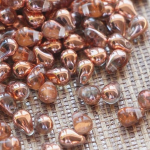 Op de afbeelding: Een verzameling druppelvormige kralen met een koperkleurige metallic afwerking. De kralen liggen verspreid over een getextureerd, lichtbruin oppervlak. Ideaal voor het maken van sieraden en knutselen.