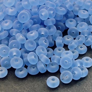 Op de afbeelding: Een stapel lichtblauwe, donutvormige glazen kralen. De kralen hebben een matte uitstraling en een gat in het midden. Deze kralen zijn geschikt voor het maken van sieraden en knutselprojecten.