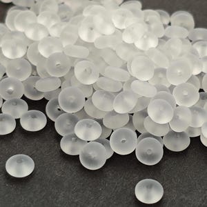 Peut inclure: Gros plan sur un tas de perles rondes translucides, percées d'un petit trou central. Les perles, de couleur blanc givré, sont disposées sur une surface sombre. Les perles sont de taille et de forme uniformes.