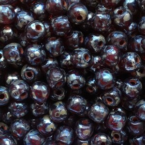 May include: A close-up shot of a pile of dark, glossy beads. The beads are a deep, iridescent color, with hints of purple and silver. Each bead has a hole running through the center, and the surface reflects light, creating a shimmering effect. These beads are suitable for jewelry making.