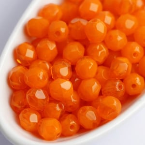 May include: A collection of faceted, orange beads displayed in a white, curved dish. The beads have a glossy, transparent appearance and are evenly distributed. The beads are a vibrant, warm orange color, and the dish is white.