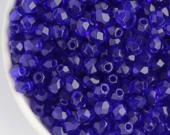 May include: A close-up shot of a white bowl filled with faceted, deep blue beads. The beads have a glossy, transparent appearance and are densely packed together. The bowl is set against a plain white background, highlighting the vibrant color of the beads.