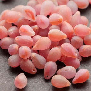 Peut inclure: Un tas de perles roses en forme de goutte d'eau, d'aspect translucide. Les perles varient du rose clair au rose plus foncé, certaines présentant des nuances orangées. Idéal pour la création de bijoux.