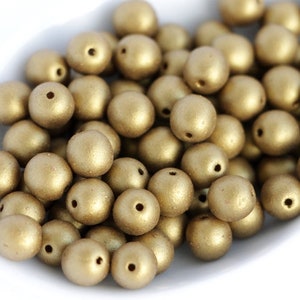 May include: A pile of gold-coloured, round beads with a matte finish. Each bead has a small hole for stringing. The beads are scattered in a white bowl, creating a textured, metallic appearance. Suitable for jewellery making and crafts.