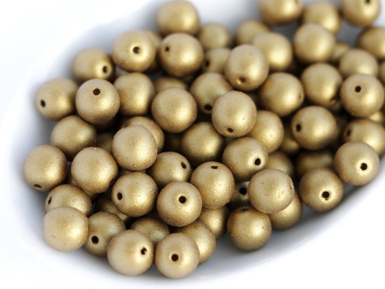 May include: A pile of gold-coloured, round beads with a matte finish. Each bead has a small hole for stringing. The beads are scattered in a white bowl, creating a textured, metallic appearance. Suitable for jewellery making and crafts.