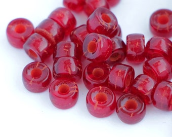 May include: A collection of small, translucent red glass beads with a cylindrical shape and a central hole. Each bead features a smooth, glossy surface with subtle internal variations and a vibrant, deep red color throughout the material.