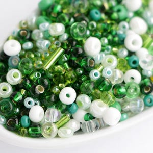 Puede incluir: Primer plano de un plato blanco lleno de una variedad de cuentas verdes y blancas. Las cuentas son en tonos esmeralda, lima y verde azulado, con algunas opciones claras y opacas. Las cuentas son de varias formas y tama&ntilde;os.