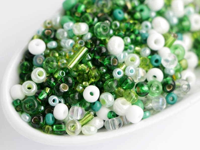 Puede incluir: Primer plano de un plato blanco lleno de una variedad de cuentas verdes y blancas. Las cuentas son en tonos esmeralda, lima y verde azulado, con algunas opciones claras y opacas. Las cuentas son de varias formas y tama&ntilde;os.