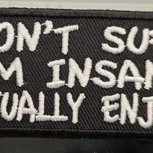 May include: Black rectangular patch with white embroidered text: "I DON'T SUFFER FROM INSANITY I ACTUALLY ENJOY IT." The patch has a black border and is likely made of fabric. The text is in a bold, sans-serif font.