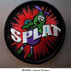 May include: A black and red embroidered patch with a green creature with a white, purple, and blue body. The creature is surrounded by a red and white explosion and the word "SPLAT" is written in white.